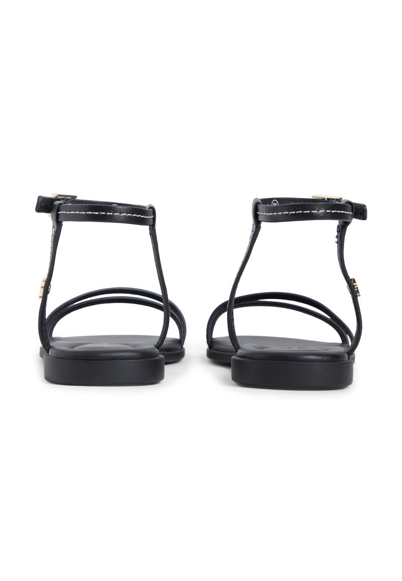 Par de sandalias planas negras con finas correas en el tobillo y los dedos, costuras visibles y pequeñas hebillas doradas, vistas desde atrás.