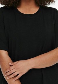 Svart kortärmad t-shirt i mjukt material. Rund halsringning och avslappnad passform. Armen korsad över kroppen, vilket framhäver textur och form.