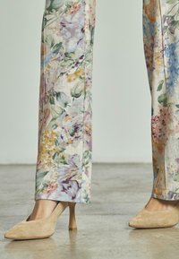 Benen in bloemenpatronen broek gecombineerd met beige suède pumps met spitse neuzen op een gepolijste betonvloer.