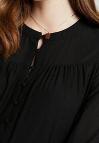 Černá blůza s kulatým výstřihem, předním zapínáním na knoflíky potažené látkou a jemným nařasením. Zlatý řetízek s obdélníkovým přívěskem.