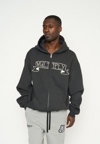 Sudadera con cremallera de color gris oscuro con un gran gráfico en blanco que dice "MULTIPLICAR" en el pecho. Presenta una capucha amplia y bolsillos frontales. Combinada con pantalones de chándal grises.