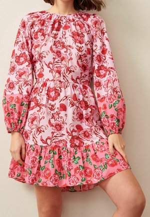 Femme portant une robe rose à fleurs avec des manches longues bouffantes et un ourlet à volants, ornée de motifs de fleurs rouges et vertes.