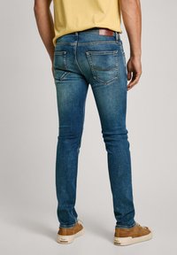 Jeans da uomo in denim blu con vestibilità slim, cinque tasche e leggera scoloritura. Presentano una toppa posteriore in pelle marrone e cuciture a contrasto.