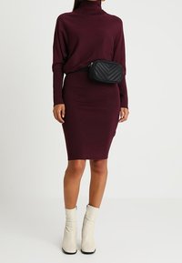 Donna che indossa un vestito a collo alto bordeaux, stivaletti bianchi alla caviglia e una borsa a tracolla nera trapuntata attorno alla vita.