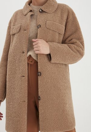 Cappotto in faux fur color tan con collo a camicia, due tasche frontali e chiusure con bottoni. Texture morbida con vestibilità rilassata, abbinato a un maglione beige.
