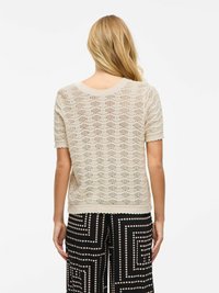 Maglione beige lavorato a maglia con motivo ondulato, maniche corte e orlo scallop, indossato sopra pantaloni neri con un design a cerchi geometrici.