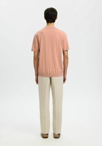 Homme debout, de dos, portant une chemise rose clair à manches courtes, un pantalon beige et des sandales marron sur un fond blanc.