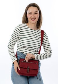 Lächelnde Frau in gestreiftem Langarmshirt und Jeans, die eine rote Umhängetasche hält und die Vordertasche mit Reißverschluss öffnet.