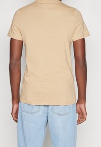 Tričko s krátkým rukávem a kulatým výstřihem z lehké béžové bavlny, přiléhavý střih s hladkou texturou a čistým šitím na ramenech.