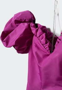 Blouse violette à manches bouffantes et col volanté, suspendue à une chaîne sur un fond uni.