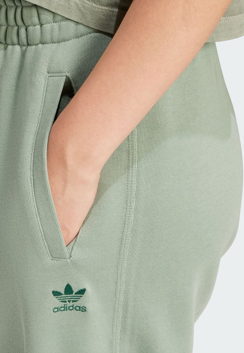 Adidas sweatpants in light green fabric feature a ribbed waistband, side pockets, and embroidered logo on the left leg. Smooth texture.