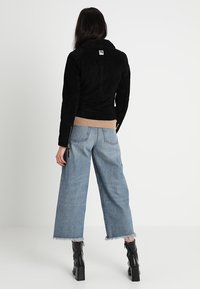 Giacca in suede nera con colletto, maniche lunghe e orlo a coste beige. Abbinata a jeans larghi blu chiaro e stivali neri alla caviglia.