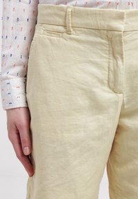 Pantalon en lin jaune pâle avec une poche latérale, une ceinture ajustée et une texture subtile. Une main repose sur le tissu, montrant sa chute.