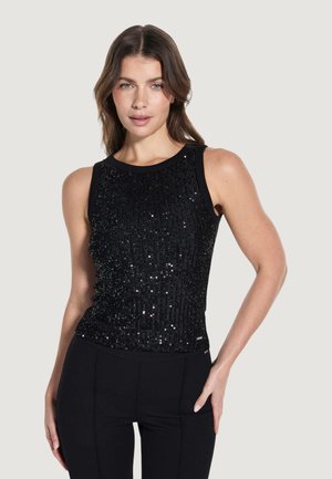 Kobieta o brązowych włosach do ramion, ubrana w czarny, bezrękawnikowy top z cekinami i czarne spodnie, pozująca z jedną ręką na biodrze.