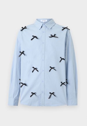 Světlomodrá košile na knoflíky s dlouhými rukávy, ozdobená mnoha malými černými mašličkami rozptýlenými po přední straně a rukávech.