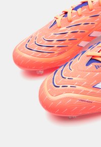 Botas de fútbol de color naranja brillante con acentos azules, parte superior sintética texturada, sistema de cordones y patrones de rayas distintivos. Tecnología HybridTouch.