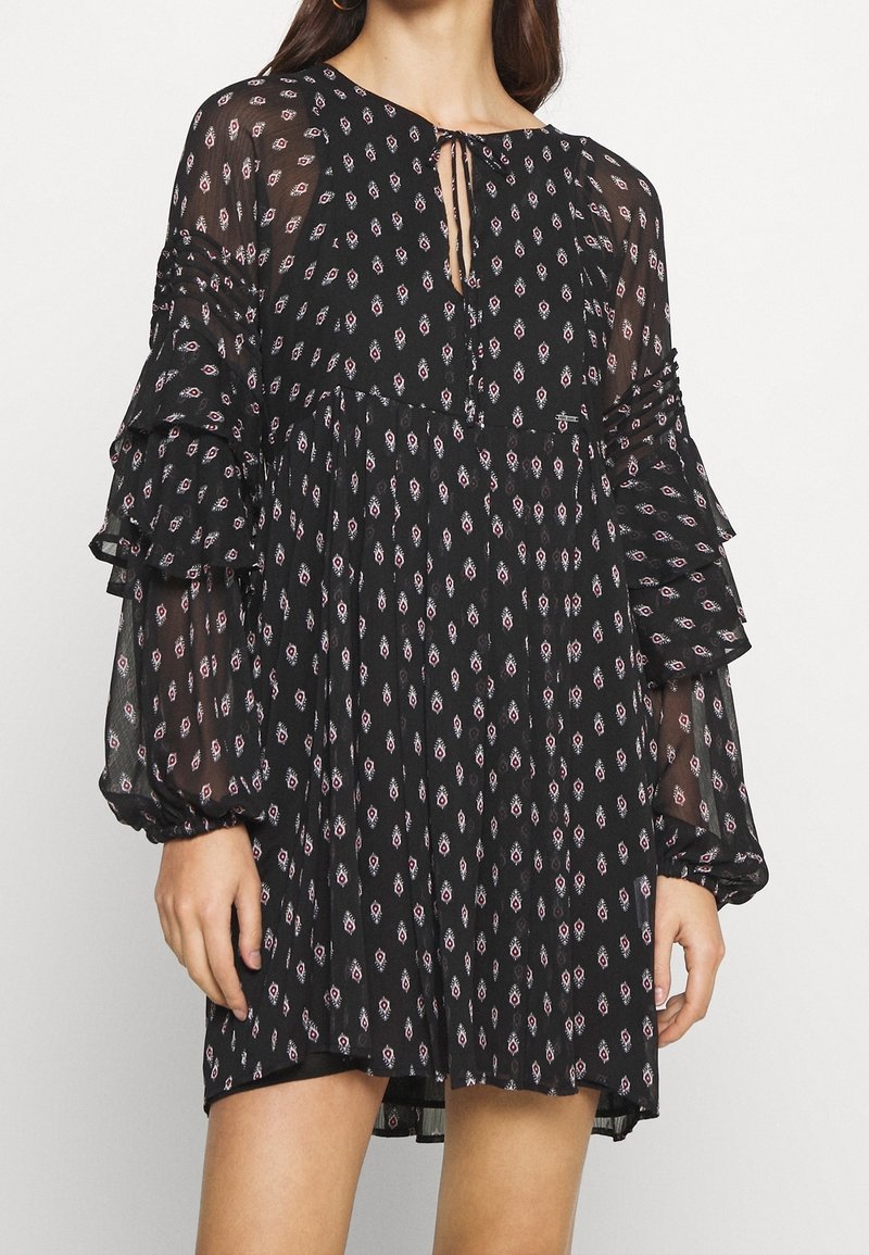 Mini-robe noire avec imprimé géométrique blanc et rouge, manches longues transparentes avec volants superposés, et petit décolleté à œillet noué à l'avant.