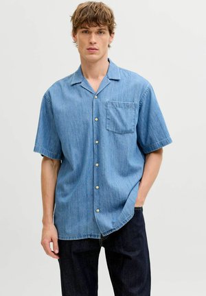 Giovane uomo che indossa una camicia di jeans leggera a maniche corte con taschino sul petto, in piedi con una mano nella tasca dei pantaloni scuri su sfondo neutro.