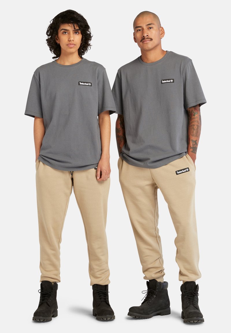 T-shirt in cotone grigio con patch logo; abbinato a pantaloni della tuta beige e stivaletti neri con lacci. Texture liscia, vestibilità rilassata.