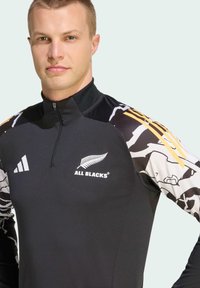 Černý sportovní top s vysokým límcem, předním zipem a bílými/kamerovými rukávy s žlutými akcenty. Zobrazuje logo "ALL BLACKS" a insignii Adidas.