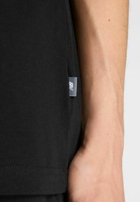 T-shirt en coton noir avec une étiquette latérale, manches courtes et texture lisse. Le tissu semble léger et ajusté.