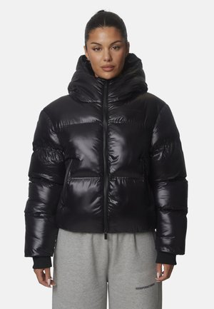 Donna con una giacca a vento nera lucida con cappuccio e pantaloni della tuta grigi, in piedi su sfondo bianco.