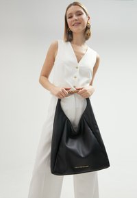 Sac hobo en cuir noir avec un design souple, doté d'un logo doré en bas et d'une surface douce et texturée.