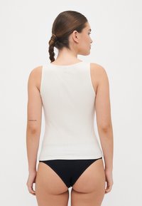 Balta šonkaidžių marškinėlių palaidinė su apvaliu iškirpimu, prigludusio silueto ir plačiomis petnešomis, dėvima su juodais bikini apatiniais.