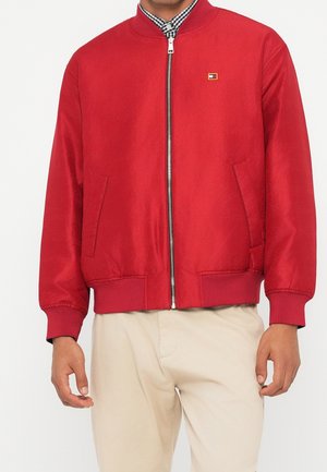 Homme portant une veste bomber rouge avec une fermeture éclair frontale, des poignets et un bas côtelés, par-dessus une chemise à carreaux et un pantalon beige.