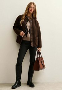 Chaqueta de forro marrón con cierre de botón, suéter argyle beige y negro, pantalones negros, botas altísimas negras y bolso marrón.