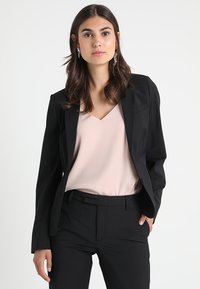 Černý blazer a kalhoty nošené přes světle růžové tričko. Blazer má strukturovaný design s vypodloženými ramenními popruhy a předními kapsami.
