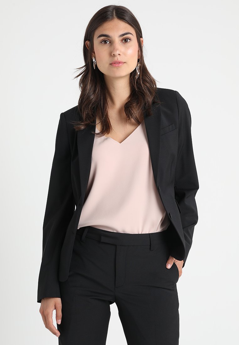 Černý blazer a kalhoty nošené přes světle růžové tričko. Blazer má strukturovaný design s vypodloženými ramenními popruhy a předními kapsami.