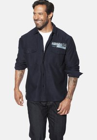 Marineblaues Hemd mit Knöpfen und zwei Brusttaschen, mit einem grafischen Druck auf der linken Seite. Kombiniert mit dunklen Jeans.