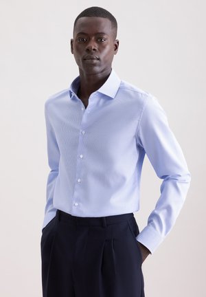 Mand iført en lyseblå skjorte med knapper, stoppet ned i mørke, plisserede bukser, stående med hænderne i lommerne mod en ensfarvet baggrund.