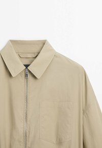 Beige collared jacket with a front zip closure and a single chest pocket. Made of lightweight material with a smooth texture.