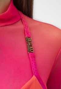 Ružičasta prozirna tkanina odjeće s naramenicom koja ima zlatni detalj logotipa Versace Jeans Couture na ramenu osobe.