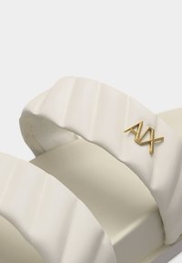 Sangles de sandales texturées blanches avec un détail logo AVA en métal doré sur la sangle supérieure, sur un fond uni.