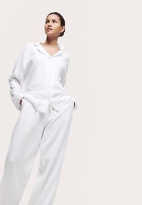 Hvid hørskjorte og bukser med vide ben, med knapper foran og lommer. Blød tekstur og afslappet pasform, mod en lys baggrund.