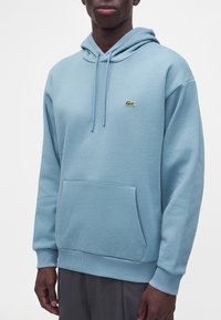 Homme portant un sweat à capuche bleu clair avec poche avant et petit logo crocodile vert sur la poitrine sur fond uni.