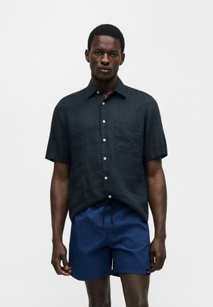 Muž oblečený v tmavej krátkosleevej košeli na gombíky a námorníckych šortkách so šnúrkou na svetlom jednoliatom pozadí.