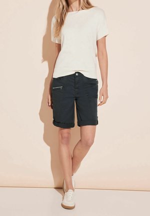 Femme portant un haut blanc à manches courtes, un short noir à mi-cuisse avec poches zippées, et des baskets blanches, se tenant contre un mur beige.