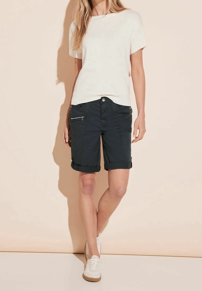 Žena v bílém krátkém rukávu, černých kraťasech ke kolenům s kapsou na zip a bílých teniskách stojící proti béžové zdi.