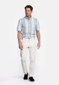 Chemise à manches courtes à rayures bleues, vertes et blanches, associée à un pantalon blanc et des chaussures noires à enfiler.
