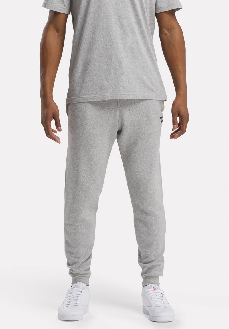 Grijze katoenen shirt met korte mouwen en bijpassende joggingbroek. De broek heeft elastische boorden en een klein zwart logo op de linker bovenbeen. Witte sneakers.