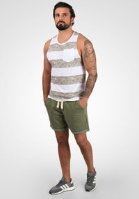 Un débardeur rayé blanc et beige avec une poche, associé à un short olive. Le modèle porte des chaussures de sport grises.