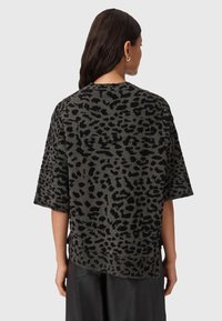 Vrouw met donker haar, gekleed in een los, kort mouwend zwart en grijs luipaardprint bovenstuk, gezien van achteren tegen een effen achtergrond.