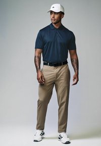 Marine mønstret polo t-shirt, beige bukser, hvid kasket, sort bælte og hvide sneakers med sorte striber. Model står foran en lys grå baggrund.