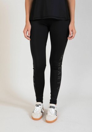 Czarne legginsy o dopasowanym kroju, z gładką fakturą i graficznym nadrukiem wzdłuż lewej nogawki. Połączone z białymi butami sportowymi.