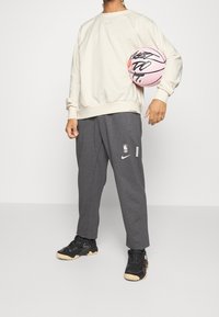 Lys beige sweatshirt med ribbet kant, grå sweatpants med NBA-logo og sorte sneakers med strukturerede såler, holder en pink basketball.