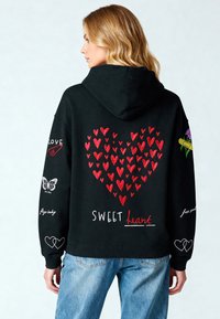 Sudadera negra con patrones de corazones rojos, texto "SWEET heart", mariposas y detalles florales. Material suave con un ajuste relajado.
